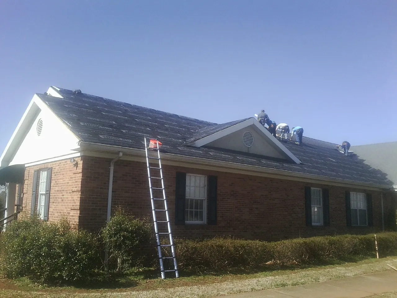 Crew tearing shingles off a brick ranch roof with a ladder set — Metal Roof Repair work by our crew in Snow Camp