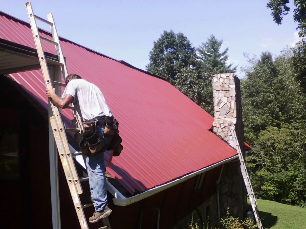 Roofer on a ladder beside a red metal roof and stone chimney — Soffit & Fascia Repair by Roofing Brothers in Snow Camp